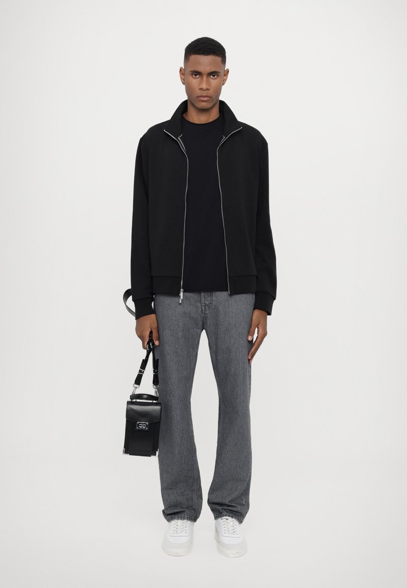 Giacca nera con zip e cuciture a contrasto, maglietta nera, jeans grigi, sneakers bianche, mentre tiene una piccola borsa nera con tracolla.