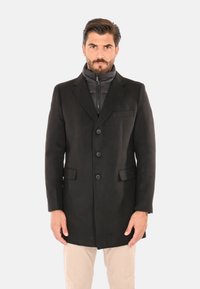Uomo con cappotto nero abbottonato sopra una giacca nera con cerniera, pantaloni beige, in piedi davanti a uno sfondo chiaro e uniforme.