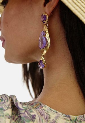 Perfil lateral de una persona que lleva pendientes colgantes de oro con tres grandes piedras preciosas facetadas de color púrpura y un top estampado en colores claros.