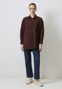 Bordeauxfärgad skjorta med knappstängning fram, långa ärmar och klassisk krage, matchad med blå denimjeans och beige sneakers.