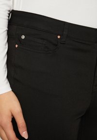 Pantalon en denim noir avec une texture lisse, doté d'une poche avant, d'un petit bouton en métal et de détails en cuivre le long de la taille.