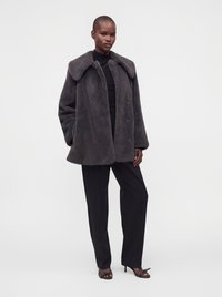 Femme portant un manteau en fausse fourrure gris foncé, un pantalon noir, un haut noir et des talons ouverts noirs, debout devant un fond clair uni.