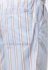 Tissu rayé bleu clair avec des rayures verticales beige et marron, comprenant une poche carrée avec un petit logo crocodile brodé.