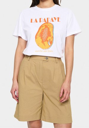 Femme portant un t-shirt blanc avec un motif papaye orange et un texte en français, associé à un short beige taille haute à hauteur des genoux.