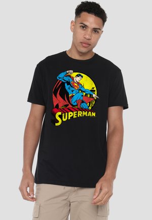 Young man wearing a black t-shirt with a vintage Superman graphic in blue, red, and yellow, paired with beige cargo shorts.