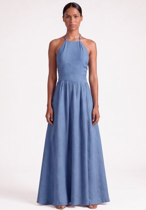 Donna che indossa un abito blu senza maniche, lungo fino a terra, con collo all'americana, in piedi davanti a uno sfondo bianco uniforme.