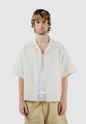 Junger Mann mit welligem blondem Haar, der ein kurzärmliges, cremefarbenes Hemd über einem weißen Tanktop und beige Hosen trägt, vor einem schlichten Hintergrund.