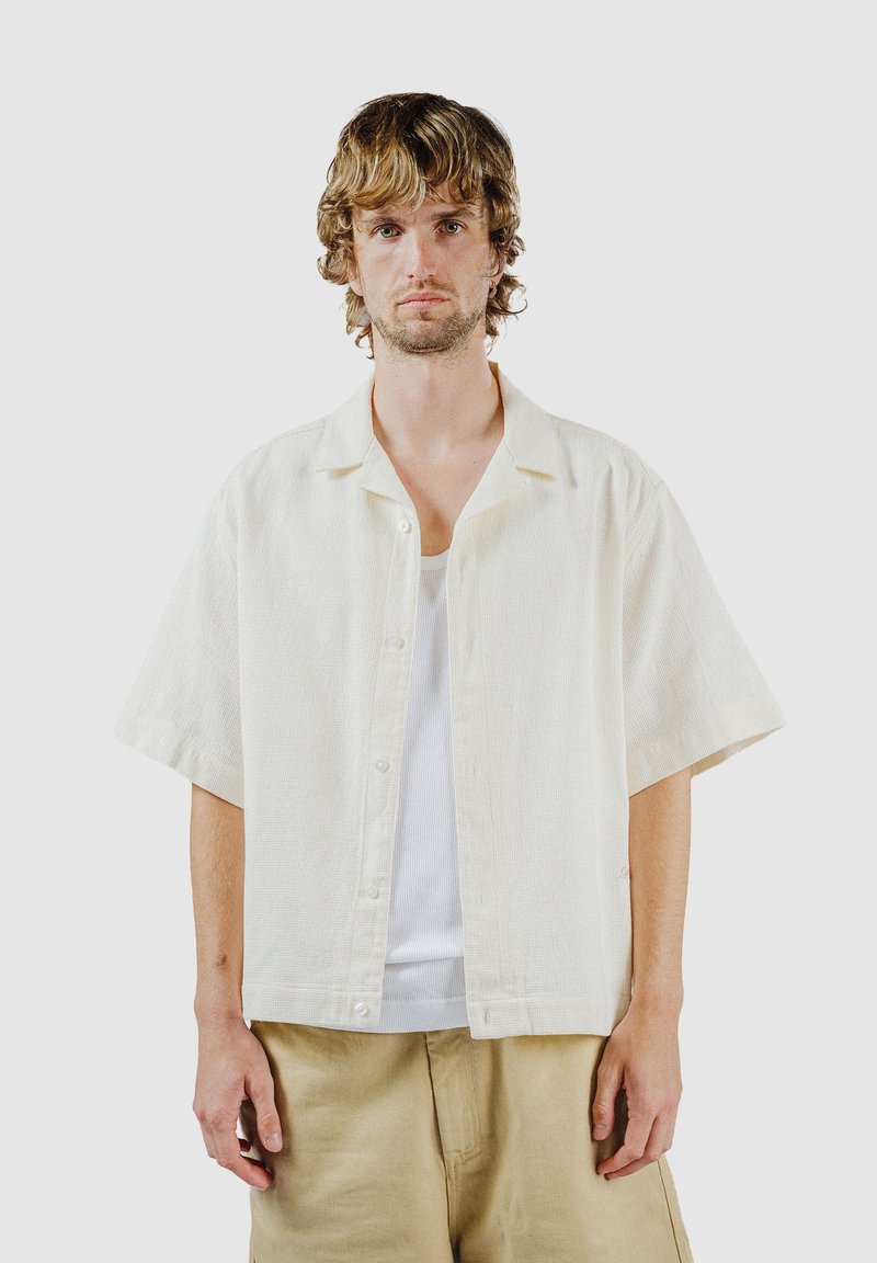 Jaunas vyras su banguotais šviesiais plaukais, dėvintis šviesios kreminės spalvos trumpomis rankovėmis marškinius su sagutėmis ant balto tanko marškinėlių ir smėlio spalvos kelnes priešais paprastą foną.