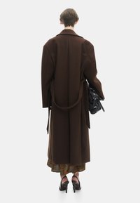 Lange bruine overjas met wijde mouwen, riemdetails en een gelaagde patroonrok eronder. Accessoires omvatten een zwarte handtas en hoge hakken.