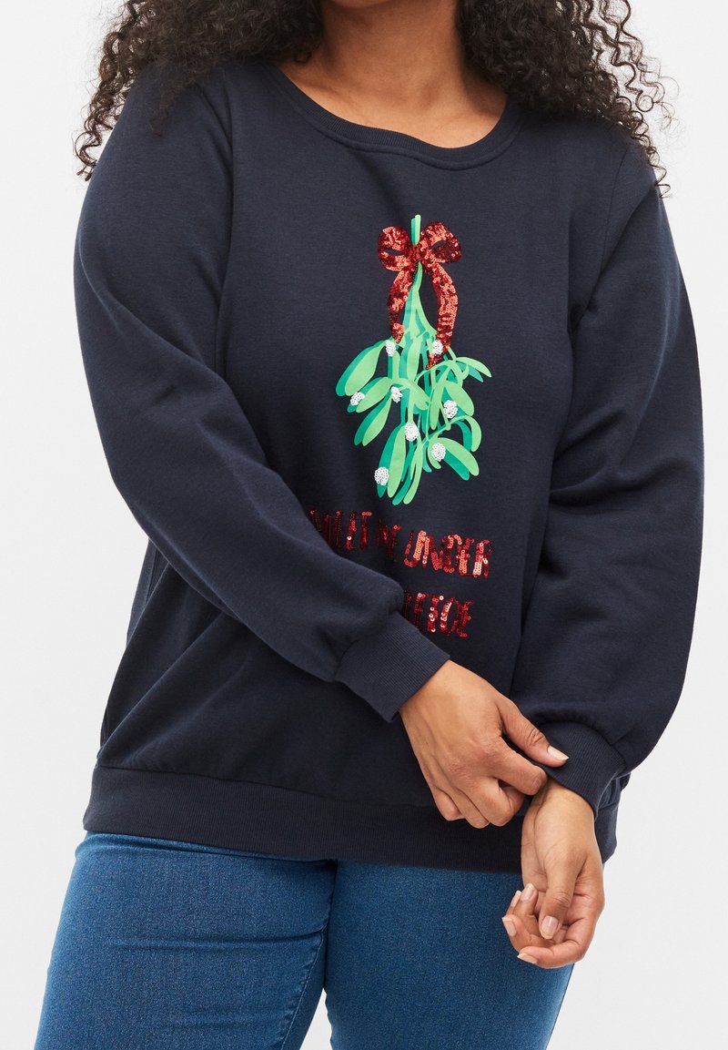 Personne portant un sweat-shirt bleu marine avec un dessin de gui vert et un nœud rouge, associé à un jean bleu, ajustant la manche.