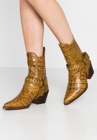 Botines de punta afilada en imitación de cocodrilo brillante de color marrón-amarillo. Tacón de altura media con correa decorativa y tachuelas. Superficie texturizada.