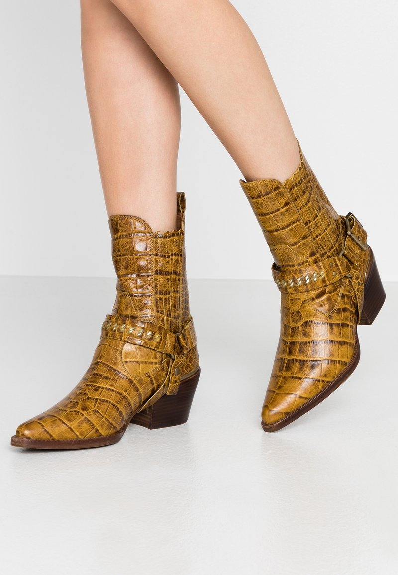 Botines de punta afilada en imitación de cocodrilo brillante de color marrón-amarillo. Tacón de altura media con correa decorativa y tachuelas. Superficie texturizada.