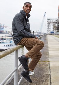 Veste imperméable noire, pantalon en velours côtelé beige, bottes de randonnée noires avec lacets, assis sur une rambarde près d'un plan d'eau. Fond industriel.