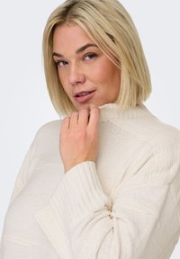 Pull en maille texturée de couleur crème avec un col montant et des poignets côtelés, présentant des rayures horizontales et une coupe décontractée.