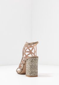 Sandal con tacco alto color beige, con cinturini e un design a taglio, decorata con accenti di gioielli. Presenta un tacco a blocco in corda intrecciata e una cinghia alla caviglia sicura.