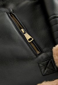 Sort lædermateriale med en gylden lynlås, med en tekstureret overflade og en fuzzy tan accent. Inkluderer en lille kviltet detalje.