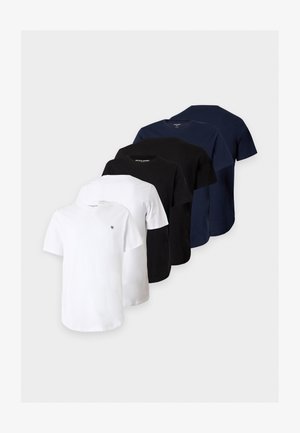 Homme debout portant un t-shirt blanc, un short large sombre jusqu’aux genoux, des chaussettes blanches et des baskets montantes noires sur un fond blanc uni.