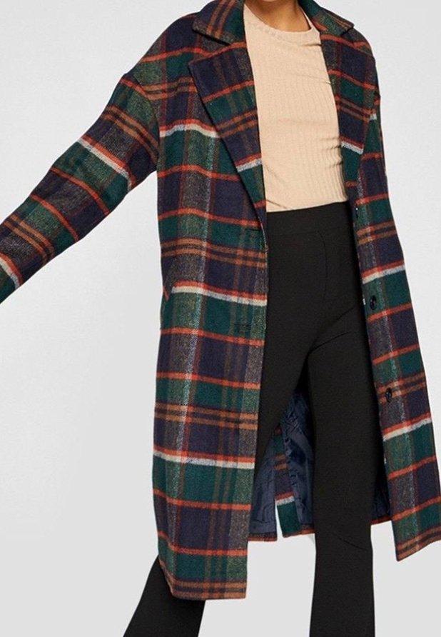 Femme portant un long manteau ouvert à carreaux avec des rayures vertes, bleu marine, orange et blanches sur un haut beige et un pantalon noir.