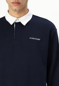 Sötétkék polo ing fehér gallérral, hímzett "GUESS JEANS" logóval. Az anyaga lágy és bordázott textúrájú.