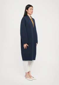 Manteau long bleu marine avec une coupe décontractée, des épaules tombantes et un design en V. Doté d'une poche avant, il est associé à un pantalon clair et des chaussures pointues.