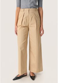 Pantaloni bej cu picioare largi, cu pliuri, talie �înaltă și țesătură moale. Asortați cu pantofi negri cu vârful ascuțit.