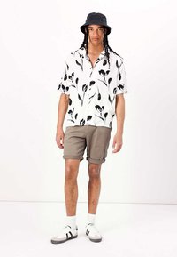 Camisa blanca de manga corta con patrón floral negro, pantalones cortos beige, sombrero de cubo negro y zapatillas blancas con rayas negras.