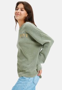 Vrouw in een groene lange mouwen shirt met de tekst "ROXY" en blauwe jeans, glimlachend, handen achter de rug, tegen een effen witte achtergrond.