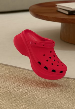 Roter Gummiclog mit Belüftungslöchern und einem hinteren Riemen, der eine abgerundete Form und eine glatte Textur zeigt, auf einer gewebten Oberfläche platziert.