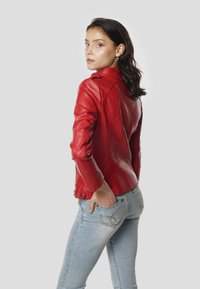 Punane nahast mootorrattajakk, millel on istuv lõige ja õlataldad. Kombineeritud siniste teksapükstega ning käsi on taga taskus.