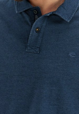 Nahaufnahme einer Person, die ein strukturiertes dunkelblaues Polohemd mit zwei Knöpfen und einem kleinen gestickten Logo auf der Brust trägt.