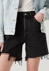 Pantalones cortos de mezclilla negra con dobladillo deshilachado, diseño de talle alto y bolsillos delanteros, combinados con una chaqueta de mezclilla azul claro.