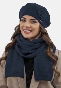 Tengerész kék kötött baret, feltekert virág részletekkel és hozzá illő sállal, puha tapintással és téglalap alakban. Meleg és kényelmes téli kiegészítők.