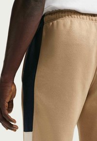 Pantalones deportivos color caqui con una franja lateral negra, que incluyen una cintura elástica y una textura suave. Se ve una mano apoyada sobre la tela.