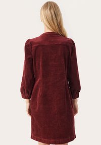 Rochie din catifea burgundă, cu o croială lejeră, mâneci bufante și decolteu rotund, având o textură ribbed și o lungime până la genunchi.
