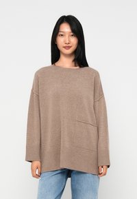 Brązowy oversize sweter z prążkowanym dołem i mankietami, posiadający kieszeń z przodu. Nosi się go na jasnoniebieskich dżinsach. Miękka tekstura i luźny krój.