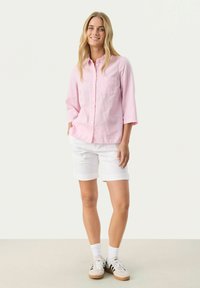 Lys pink, langærmet skjorte med knapper og brystlomme, parret med hvide shorts. Modellen har hvide sokker og sneakers på.
