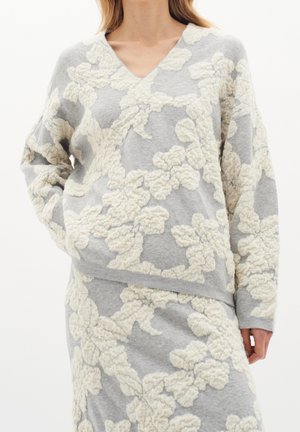 Femme portant un pull gris à col en V et une jupe assortie avec des motifs floraux texturés crème, mains dans les poches et coupe décontractée.
