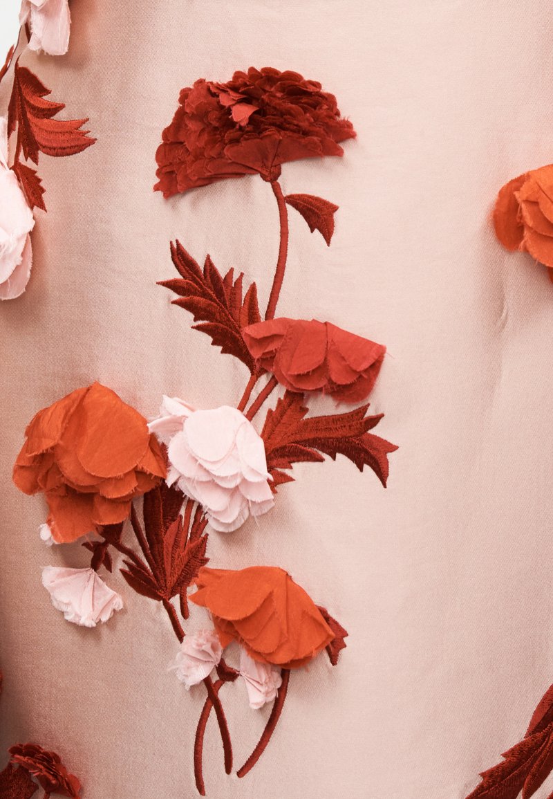 Tessuto con fiori di stoffa rossi e rosa stratificati e foglie ricamate su uno sfondo rosa chiaro.