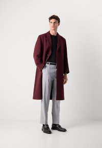 Palton din amestec de lână bordeaux, cu guler cu unghii, închidere cu un singur nasture, asortat cu pantaloni gri deschis, tăiați, și pantofi din piele negri.