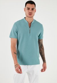 Polo majica s kratkimi rokavi in zadrgo v svetlem teal odtenku z strukturirano tkanino. Ima sproščen kroj in subtilen vzorec, nosi se z belimi hlačami.