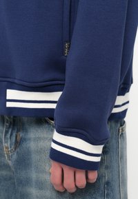 Námořnicky modrá mikina s bílými proužkovanými žebrovanými manžetami a lemem, vybavená kapsou na zip a texturovanou tkaninou. Ruka viditelná v popředí.