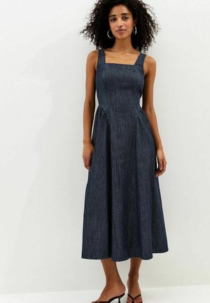 Kobieta o kręconych włosach, ubrana w bezrękawnikową, ciemnoniebieską sukienkę midi z denimu oraz czarne sandały na obcasie, stojąca na tle jasnego, gładkiego tła.