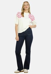 Maglione bianco a maglia con ricami floreali rosa sulle spalle, abbinato a jeans svasati blu scuro e sneakers crema. Texture liscia, design informale.