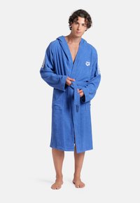 Blauwe capuchon badjas van badstof, met zakken en een strikceintuur, voorzien van een logo op de borst en patroonaccenten op de mouwen.