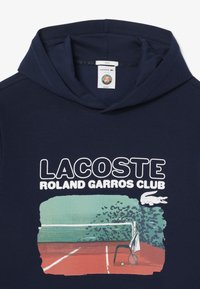 Marinblå hoodie med kenguruficka. Har texten "LACOSTE ROLAND GARROS CLUB" och en grafik av en tenniscourt i grönt och rött.