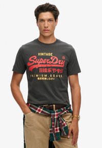 Grå bomulds-T-shirt med rød og gul "SuperDry" grafik. Båret med khaki-bukser og en grønternet skjorte bundet om livet.