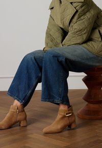 Bottes en daim couleur marron clair à talon bloc, fermeture éclair latérale et détail boucle. Portées avec un jean large bleu foncé et une veste matelassée vert olive.