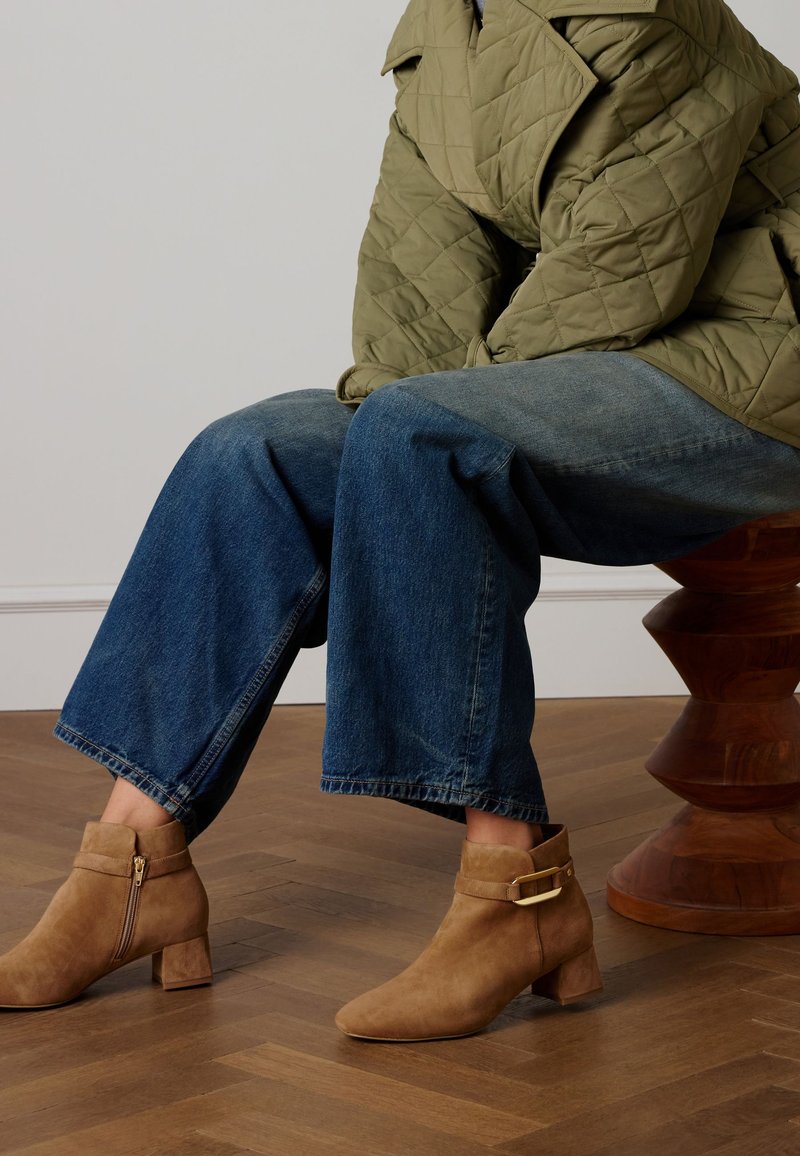 Bottes en daim couleur marron clair à talon bloc, fermeture éclair latérale et détail boucle. Portées avec un jean large bleu foncé et une veste matelassée vert olive.