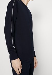 Personne portant un pull à manches longues bleu marine à col en V avec une bande blanche le long du bras et un pantalon bleu marine assorti.