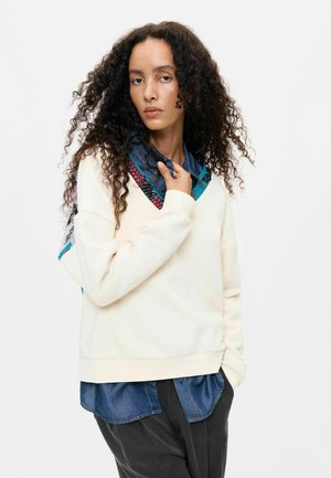 Frau mit lockigem Haar trägt einen cremefarbenen Pullover über einem blauen Hemd mit Knöpfen und dunklen Hosen, posiert mit einer Hand, die den Pulloverkragen hält.